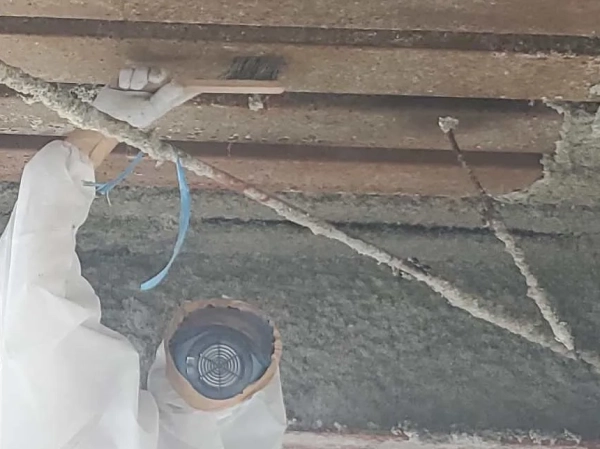 防塵マスクと防護服に身を包んだ株式会社エイキの従業員が、ブラシで鉄骨のアスベストを擦りながら落としている。