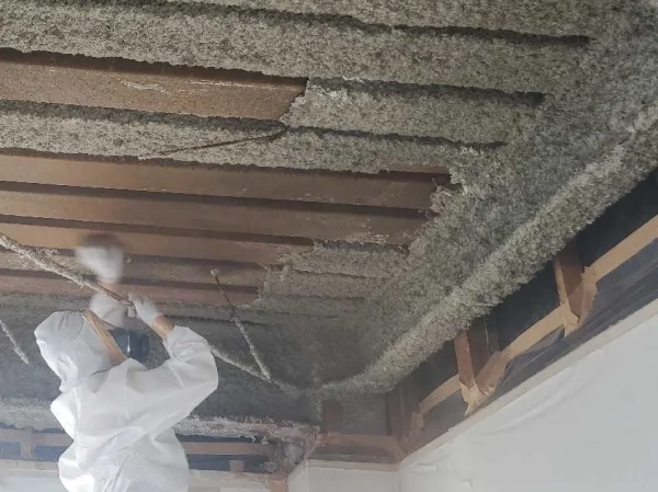 防塵マスクと防護服に身を包んだ株式会社エイキの従業員が、タワシで天井鉄骨にこびり付いたアスベストを落としている。