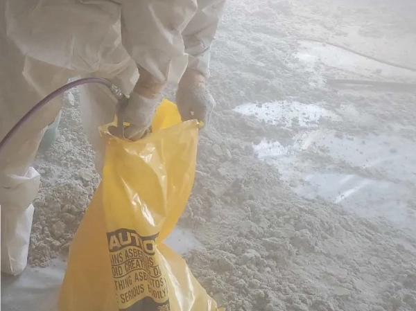 防護服に身を包んだ株式会社エイキの従業員が、除去済みアスベストを詰めた黄色の袋に飛散対策として、飛散防止剤を噴霧器で吹き付けている。