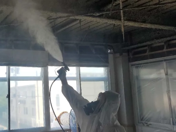 防塵マスクと防護服に身を包んだ株式会社エイキの従業員が、噴霧器でアスベスト飛散防止剤を吹き付けている。