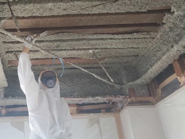 防塵マスクと防護服に身を包んだ株式会社エイキの従業員が、ケレンで湿潤化済みの吹き付け材アスベストを落としている。