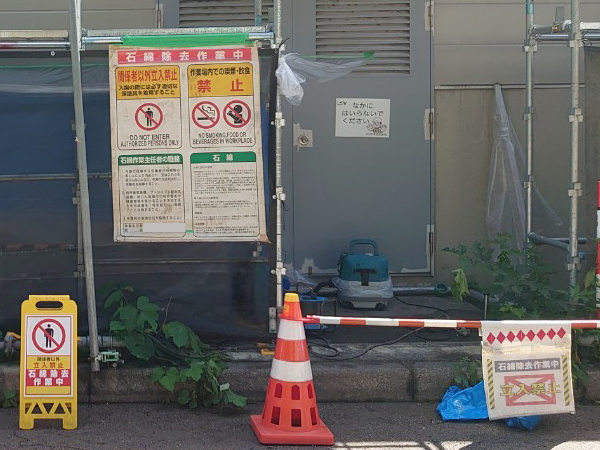 建物の扉の前に、アスベスト除去を示す表示看板と、ロードコーンがある。