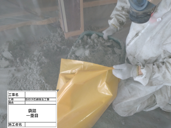 作業員が、床に落ちた吹き付けアスベストを塵取りで回収し、廃棄袋に入れている。