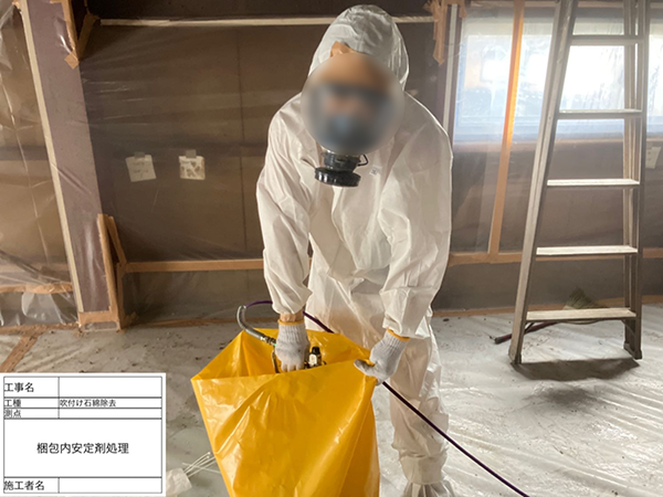 作業員が、吹き付けアスベストの入った廃棄袋の中に薬剤を散布している。