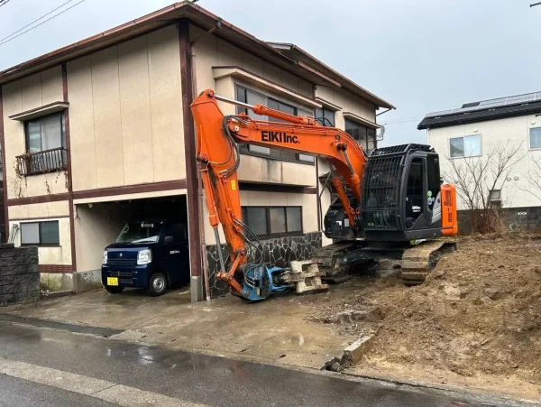 富山市の木造2階建て解体工事現場。株式会社エイキのオレンジ色の重機が投入され、解体予定の木造2階建てと並んでいる