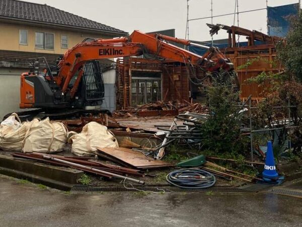 株式会社エイキのオレンジ色の油圧ショベルが木造2階建ての住宅を解体中。左手前には廃材を詰め込むフレコンバックが3つ陳列されている