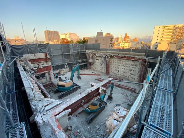 解体するビルの屋上。2台の小型重機が解体作業を進めており、途中まで切断された鉄骨がある。周囲の街並みが写っている。