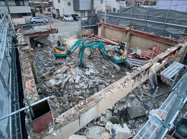 建物が解体され、地表の基礎コンクリートに挑む重機の姿が二台。養生シートは、まだそのまま被われている。
