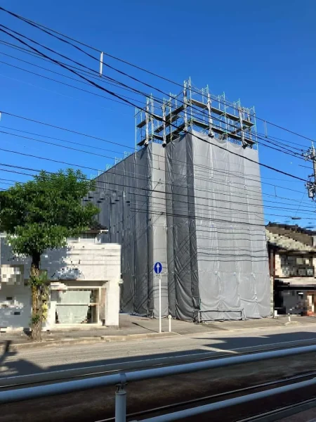 ビルの周囲をシートで囲ってある現場ビル。前面は道路に面している。隣にも建物がある。