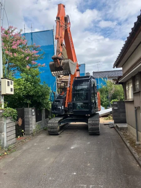 解体現場につながる前道に重機（油圧ショベル）が見えます。油圧ショベルの右側には隣家、左側には門と塀が見えます。