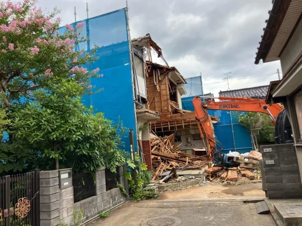 住宅を油圧ショベルで解体しています。住宅を囲むように青色のシートが見えます。