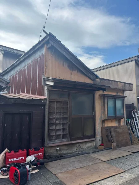 富山県内の街中にある古いRC造の納屋。経年劣化したトタン壁が目立ちます。前道を傷つけないよう板で養生されています。