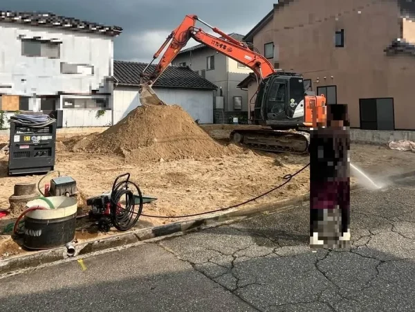 整地に向けて、油圧ショベルが土地を整えている。また株式会社エイキの作業員が散水をしている。