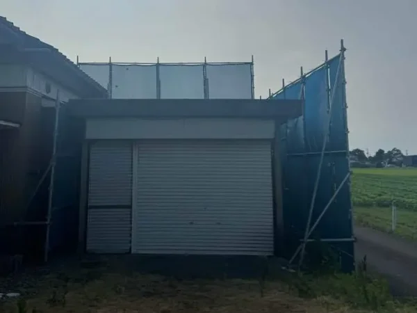 納屋の現場写真。左に家、右に田圃が見えている。納屋の周囲を養生している。