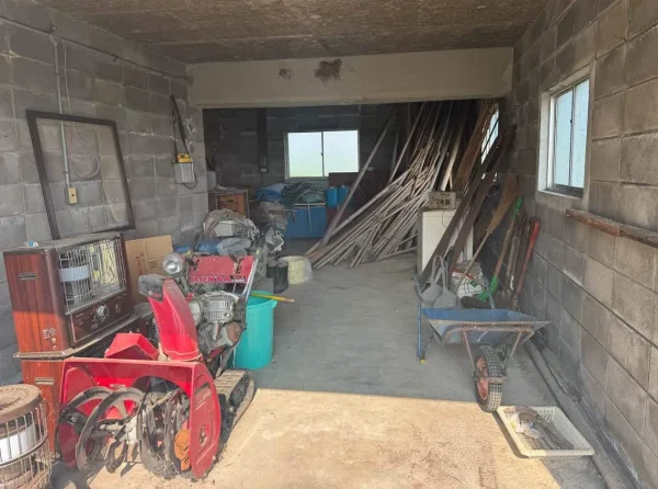 解体前の納屋の中の写真。壁はコンクリート製で、赤色の除雪機や茶色のストーブが埃をかぶっている