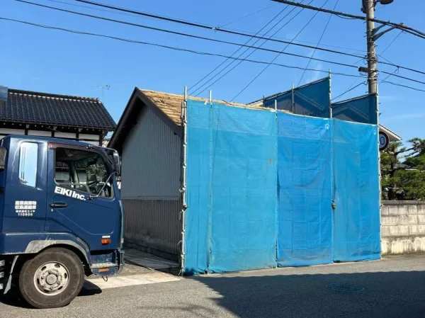 納屋の周囲（隣家と前道側）を養生してある写真。青色シートが見える。左側には、エイキの廃棄物収集運搬車が待機中。