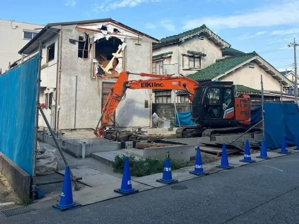 株式会社エイキのロゴが入った重機が、現場住宅を屋外から解体中。住宅の外装に穴が開いている