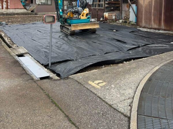 更地の上に黒色のシートが覆っている。その上に重機が一台。これから砕石をまく。