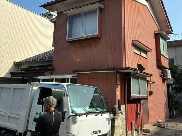 現場の解体前の住宅の写真。隣の建物のに住宅が接している。茶色の建物の前に白いトラックが停まっている。