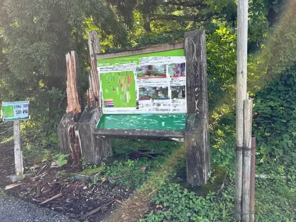 山道の途中に古びた案内板が建っている。案内板を支える左右の支え木は経年によって壊れている。