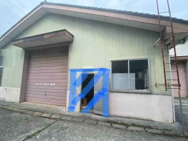 倉庫の外観が写っている。荷物の出し入れ口が狭いのが分かる。その出入り口の写真に青色マーカーで、今回工事をする拡充部分を描いた写真。