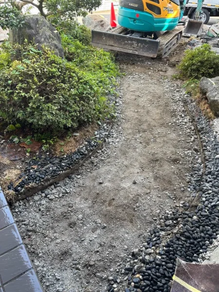 通路部分が剥がされ、砂利が見えている状態の写真。奥には油圧ショベルが見える。