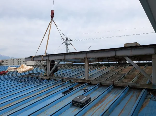 鉄骨の架台に玉掛け作業をしている写真。青い屋根の上の鉄骨の架台とクレーンの先端をロープで繋いでいる