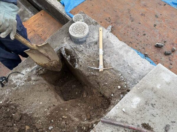 排水桝のそばのコンクリートを手作業で斫っている。ショベルで地中の土を撤去している。