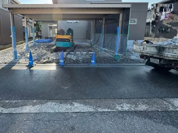 カーポートの下に重機が入り、土間を斫っている写真。右側には、不要物を積み込むトラックが待機している。