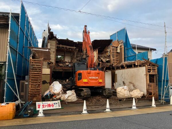 本体の住宅を重機で解体中。左右には足場とシートが見える。前面道路には、エイキのコーンが立ててある。