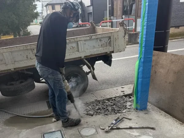 職人がコンクリート土間をブレーカーで斫っている。地面にある水道のマス（点検口）の周りには、あらかじめ四角く切れ目を入れてある。