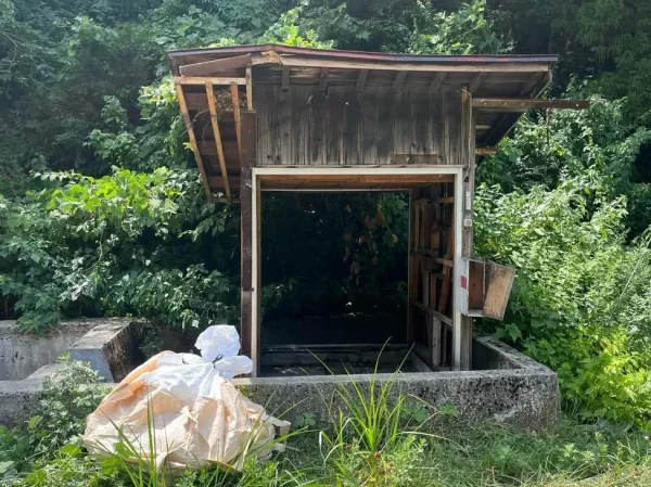 小屋・納屋。頑丈そうなコンクリート基礎が見える。奥はうっそうとした緑。