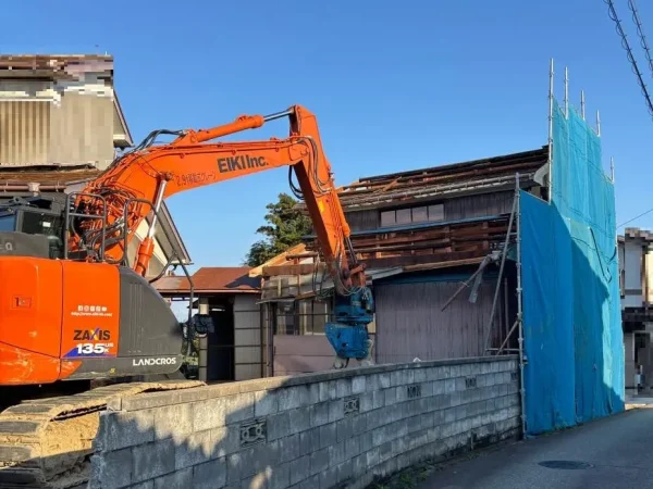 重機で、構造体を解体中。お隣の敷地や道路に面した場所には足場と青い養生シートを張っている。