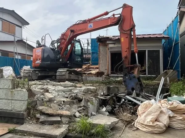 オレンジ色の重機が住宅を解体中。写真右側に半分ほど壊された住宅、写真右下側には廃材を入れるためのトンパックが置いてある。